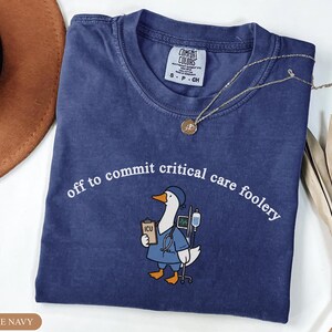 May include: A true navy blue short-sleeve t-shirt featuring a graphic of a goose dressed as a medical professional with a stethoscope, clipboard labeled ICU, and IV pole. The text reads off to commit critical care foolery in white lettering.