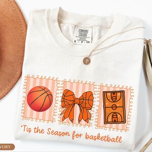 Peut inclure: T-shirt ivoire avec un motif sur le th&egrave;me du basketball. Le motif comprend un ballon de basket, un n&oelig;ud fait de ballons de basket et une illustration d'un terrain de basket. Le texte dit : "'Tis the Season for basketball."