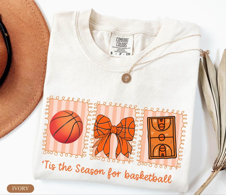 Peut inclure: T-shirt ivoire avec un motif sur le th&egrave;me du basketball. Le motif comprend un ballon de basket, un n&oelig;ud fait de ballons de basket et une illustration d'un terrain de basket. Le texte dit : "'Tis the Season for basketball."