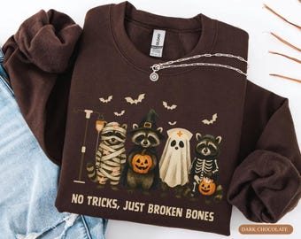 May include: A dark chocolate brown crewneck sweatshirt featuring a graphic of four raccoons dressed in Halloween costumes including a mummy, a witch, a ghost, and a skeleton, with the text NO TRICKS, JUST BROKEN BONES printed below.