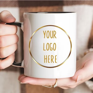 May include: A white ceramic mug with a black handle and interior, featuring a gold circular frame on the front containing the text YOUR LOGO HERE in gold lettering. The mug is held by two hands, showcasing the design clearly against a neutral background.