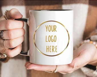 May include: A white ceramic mug with a black handle and interior, featuring a gold circular frame on the front containing the text YOUR LOGO HERE in gold lettering. The mug is held by two hands, showcasing the design clearly against a neutral background.
