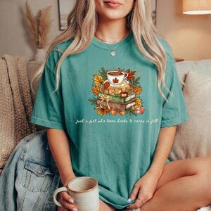 May include: A teal short-sleeve t-shirt featuring a graphic of a coffee mug, stacked books, and autumn leaves. The text "just a girl who loves books & cocoa in fall" is printed below the illustration in a white cursive font.