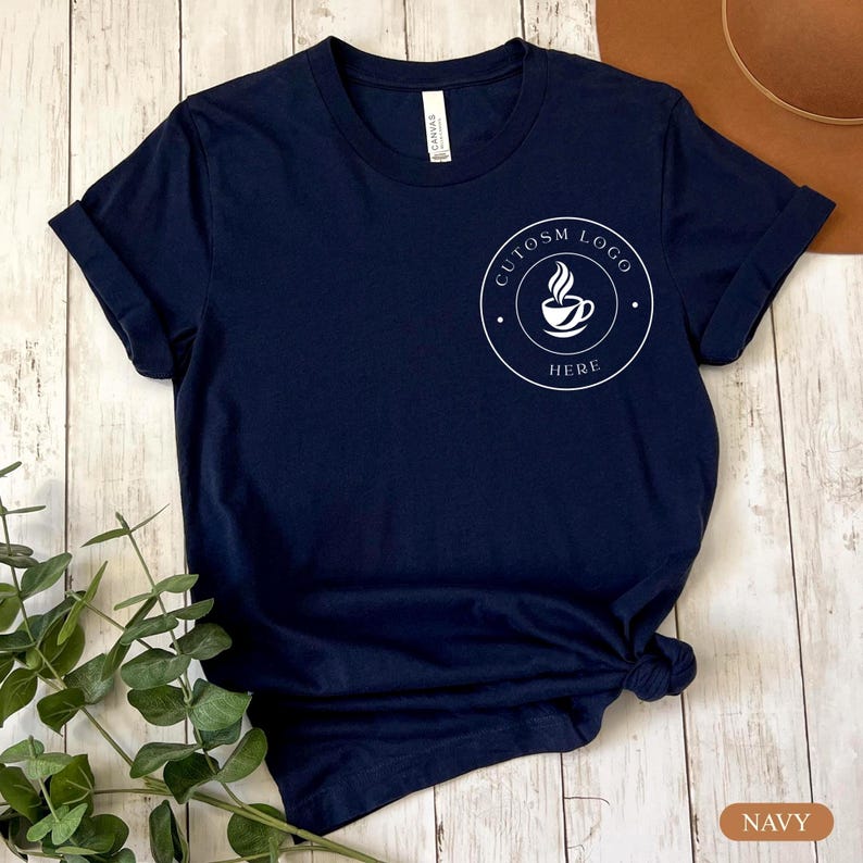 May include: A navy blue short-sleeve t-shirt featuring a white circular graphic on the left chest. The graphic contains a stylized coffee cup icon with steam and the text "CUTOSM LOGO HERE" written in a circular border around the cup.