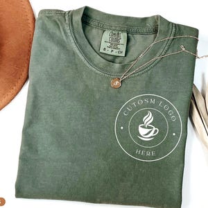May include: A moss green short-sleeve t-shirt featuring a white circular graphic on the chest with a coffee cup icon and the text CUTOSM LOGO HERE. The shirt has a Comfort Colors brand label at the inner neckline and a thin gold-toned necklace.