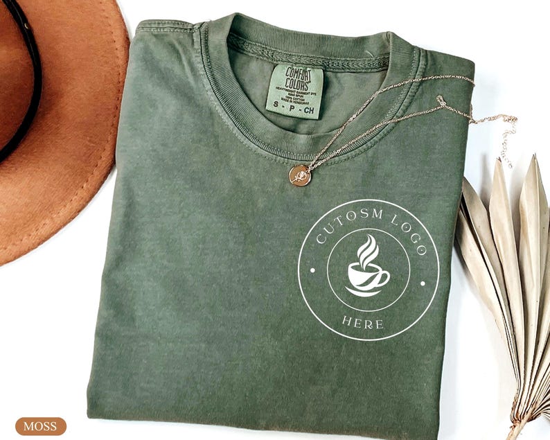 May include: A moss green short-sleeve t-shirt featuring a white circular graphic on the chest with a coffee cup icon and the text CUTOSM LOGO HERE. The shirt has a Comfort Colors brand label at the inner neckline and a thin gold-toned necklace.