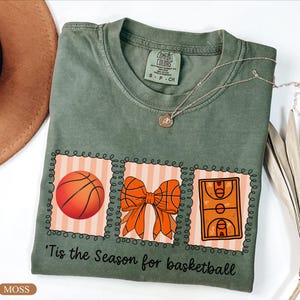 Peut inclure: T-shirt vert mousse avec un motif sur le th&egrave;me du basketball. Le motif comprend un ballon de basket, un n&oelig;ud et une illustration d'un terrain de basket. Le texte "'Tis the Season for basketball" est imprim&eacute; en dessous.