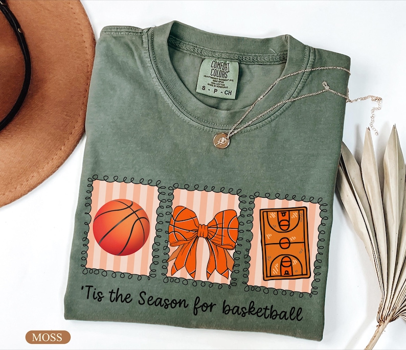 Peut inclure: T-shirt vert mousse avec un motif sur le th&egrave;me du basketball. Le motif comprend un ballon de basket, un n&oelig;ud et une illustration d'un terrain de basket. Le texte "'Tis the Season for basketball" est imprim&eacute; en dessous.