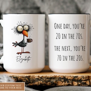 May include: White ceramic mug with a black handle and rim. One side features a cartoon bird with a coffee cup, the name "Elizabeth" is below. The other side reads "One day, you're 20 in the 70s. The next, you're 70 in the 20s."