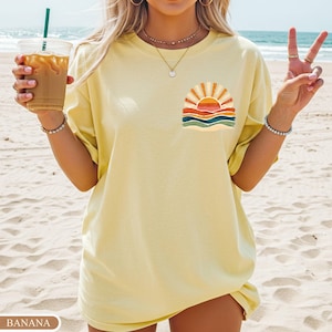 May include: A pale yellow t-shirt with a sunset design on the chest. The graphic shows a sun with orange and yellow rays, above blue and green waves. The word "BANANA" is printed on a label at the bottom.