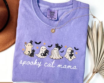 May include: A violet short-sleeved t-shirt with a graphic print featuring four white ghosts and black cats, bats, and orange stars. The text "spooky cat mama" is printed in white below the graphic. The shirt has a Comfort Colors brand label.