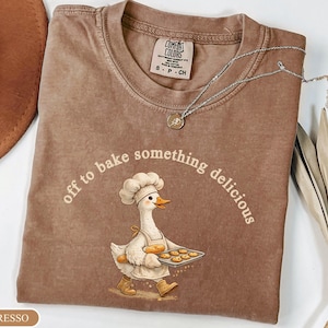 May include: An espresso-colored t-shirt featuring a graphic of a white goose wearing a chef hat and apron, holding a tray of cookies and a baguette. The text above the goose reads, off to bake something delicious. The shirt has a crew neckline.