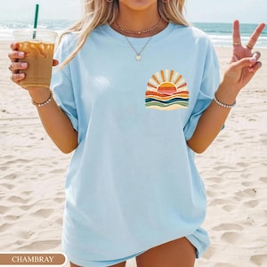 May include: Pale blue t-shirt with a sunset graphic on the front. The graphic shows a sun with orange and yellow rays, above layers of orange, red, and green waves. The shirt has a loose fit.