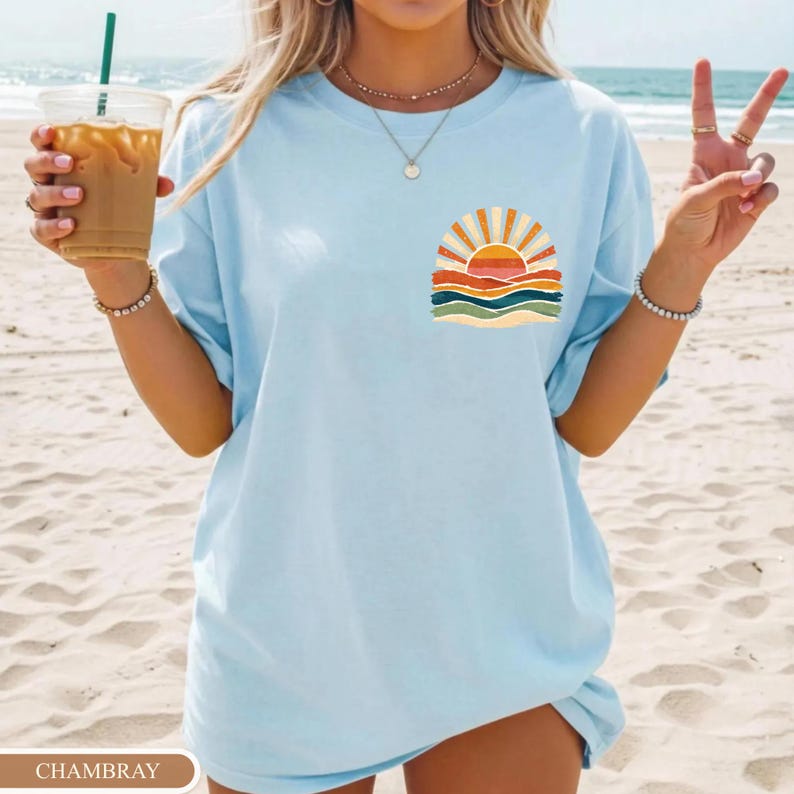 May include: Pale blue t-shirt with a sunset graphic on the front. The graphic shows a sun with orange and yellow rays, above layers of orange, red, and green waves. The shirt has a loose fit.