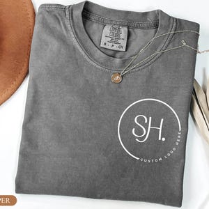 May include: A pepper-colored crew neck t-shirt featuring a white circular graphic on the chest with the letters SH and the text CUSTOM LOGO HERE. The shirt has a Comfort Colors brand label inside the collar and is displayed with a gold necklace.