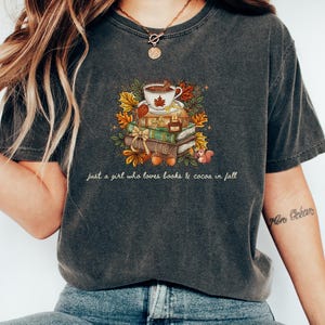 May include: A charcoal gray short-sleeve t-shirt featuring a graphic of a stack of books topped with a cup of cocoa, surrounded by autumn leaves, acorns, and a small squirrel. The text reads, just a girl who loves books & cocoa in fall.