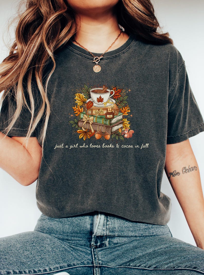May include: A charcoal gray short-sleeve t-shirt featuring a graphic of a stack of books topped with a cup of cocoa, surrounded by autumn leaves, acorns, and a small squirrel. The text reads, just a girl who loves books & cocoa in fall.