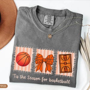 Peut inclure: Un t-shirt gris Comfort Colors avec un motif sur le th&egrave;me du basketball. Le motif comprend un ballon de basket, un n&oelig;ud et une illustration d'un terrain de basket. Le texte "'Tis the Season for basketball" est imprim&eacute; en dessous.