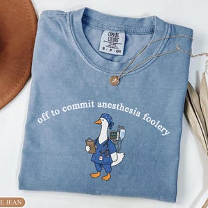 May include: A blue jean colored t-shirt featuring a graphic of a goose dressed in blue medical scrubs, a surgical cap, and a stethoscope. The goose holds a clipboard and stands near a medical monitor. The text reads, off to commit anesthesia foolery.