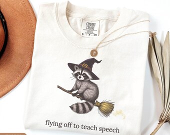 May include: An ivory t-shirt featuring a graphic of a raccoon wearing a purple witch hat and riding a broomstick. Below the graphic, the text reads "flying off to teach speech". The shirt has a Comfort Colors brand label at the inner neckline.
