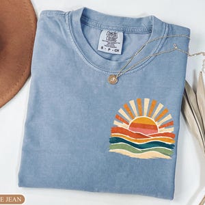 May include: A blue jean Comfort Colors t-shirt with a colourful sunset graphic. The graphic features a sun with orange and yellow rays, above layered orange, red, green, and blue waves. A silver necklace with a pendant is draped on the shirt.