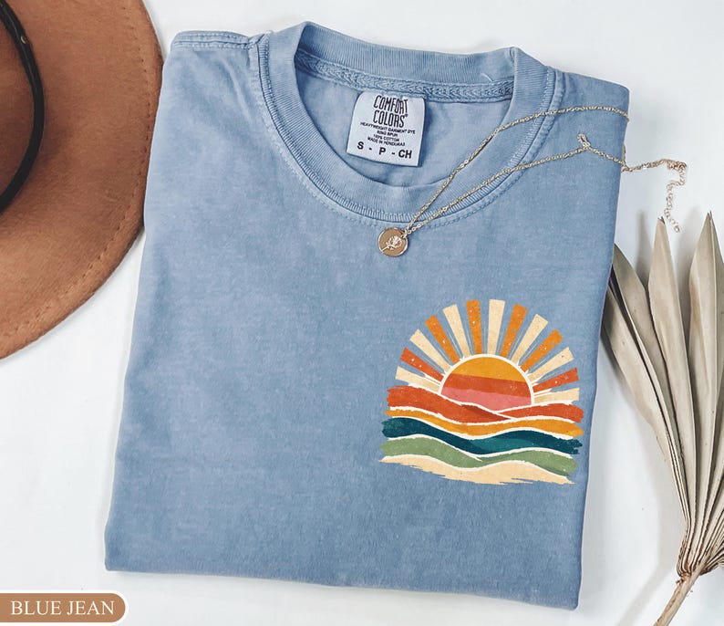 May include: A blue jean Comfort Colors t-shirt with a colourful sunset graphic. The graphic features a sun with orange and yellow rays, above layered orange, red, green, and blue waves. A silver necklace with a pendant is draped on the shirt.