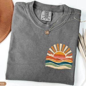 May include: A grey Comfort Colors t-shirt featuring a sunset graphic. The design shows a sun with orange and yellow rays, above layered waves in orange, red, blue, and green. A silver necklace with a small pendant is draped on the shirt.