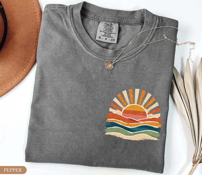 May include: A grey Comfort Colors t-shirt featuring a sunset graphic. The design shows a sun with orange and yellow rays, above layered waves in orange, red, blue, and green. A silver necklace with a small pendant is draped on the shirt.