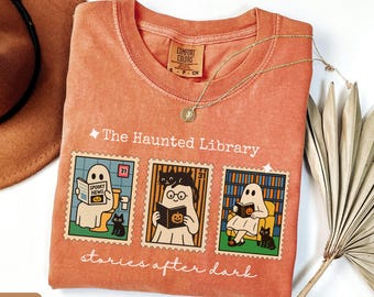 May include: A burnt orange t-shirt featuring a graphic of three postage stamps with ghosts and black cats. The text reads The Haunted Library and stories after dark. The shirt has a crew neck and a Comfort Colors label on the inside collar.