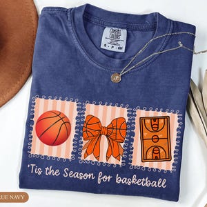 Peut inclure: T-shirt bleu marine avec un motif sur le th&egrave;me du basket-ball. Le motif comprend un ballon de basket, un n&oelig;ud et une illustration d'un terrain de basket. Le texte "'Tis the Season for basketball" est imprim&eacute; en dessous. Un collier argent&eacute; est pos&eacute; sur le t-shirt.