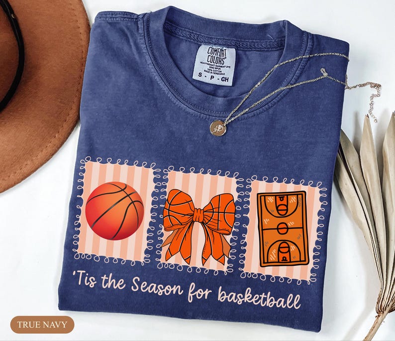 Peut inclure: T-shirt bleu marine avec un motif sur le th&egrave;me du basket-ball. Le motif comprend un ballon de basket, un n&oelig;ud et une illustration d'un terrain de basket. Le texte "'Tis the Season for basketball" est imprim&eacute; en dessous. Un collier argent&eacute; est pos&eacute; sur le t-shirt.