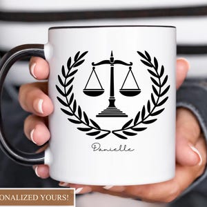 Pu&ograve; includere: Tazza in ceramica bianca con manico e bordo neri, con un emblema nero della bilancia della giustizia all'interno di una corona d'alloro. Il nome "Danielle" &egrave; scritto sotto. La tazza &egrave; tenuta da una persona.