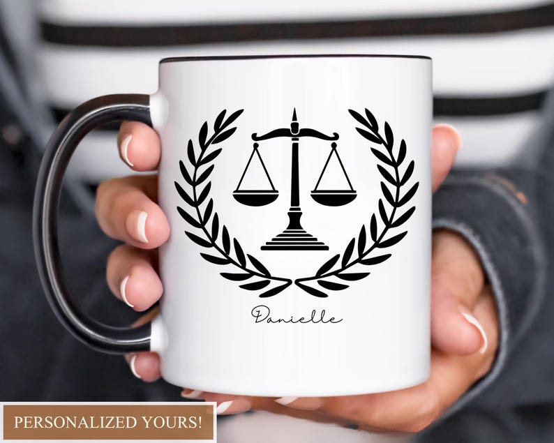 Pu&ograve; includere: Tazza in ceramica bianca con manico e bordo neri, con un emblema nero della bilancia della giustizia all'interno di una corona d'alloro. Il nome "Danielle" &egrave; scritto sotto. La tazza &egrave; tenuta da una persona.