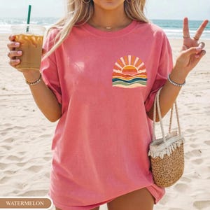May include: A pink t-shirt with a small sunset design on the chest. The graphic shows a sun with orange and yellow rays, above layers of orange, blue, and green waves. The shirt is a loose fit and the colour is watermelon.