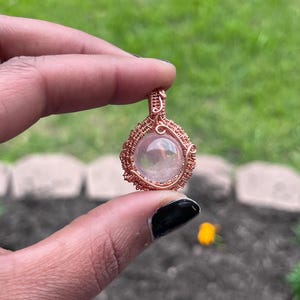 May include: A handcrafted pendant featuring a clear, round gemstone encased in intricately woven copper wire. The wirework forms a decorative cage around the stone, creating a unique and artistic piece of jewelry. The pendant is held up against a blurred green background.