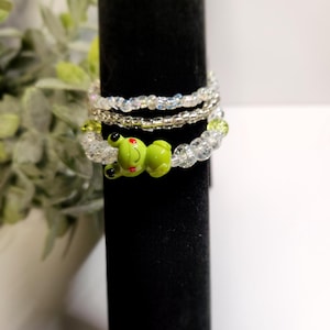 Puede incluir: Un conjunto de anillos de cuentas apilados en un expositor negro. Los anillos presentan cuentas transparentes e iridiscentes, con un anillo que muestra un dije de rana verde con detalles rojos. Un accesorio divertido y caprichoso.