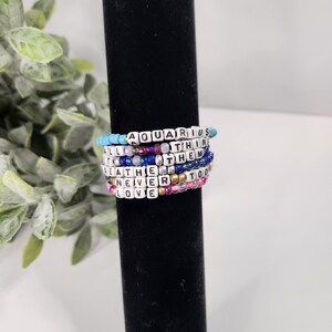 May include: A stack of colorful beaded bracelets with words spelled out on each one. The words are "Aquarius", "All", "Think", "The", "Them", "Breathe", "Never", "Too", and "Love".