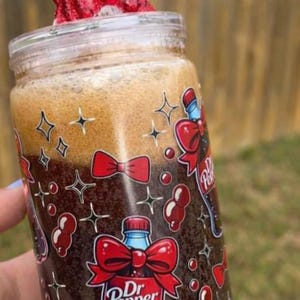 Może przedstawiać: Przezroczysty szklany słoik z motywem Dr. Pepper, wypełniony ciemnobrązowym napojem i zwieńczony pianą. Słoik jest ozdobiony czerwonymi kokardkami i logo Dr. Pepper. Czerwona kokarda zdobi pokrywkę, a włożona jest przezroczysta słomka.