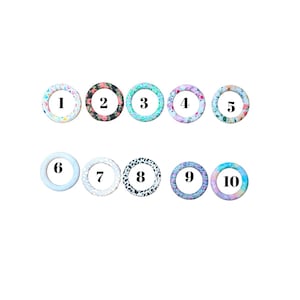 May include: Ten round silicone teething rings with various patterns. Designs include floral, geometric, and animal prints in colors like pink, blue, and black. Each ring is numbered from one to ten.