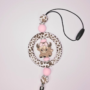 May include: A pink and brown silicone baby teether with a cow design. The teether has a ring with a cow inside and a tassel on the bottom.