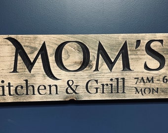 May include: Wooden sign with the words "MOM'S Kitchen & Grill" carved into the wood. The sign also includes the hours of operation, 7 AM to 6 PM, Monday through Friday. The sign is a light brown color.