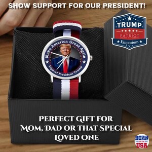 Puede incluir: Un reloj de pulsera con una correa a rayas rojas, blancas y azules y una esfera con un retrato y el texto "Make America Great Again". El reloj est&aacute; en una caja de regalo negra con el texto "Show Support For Our President!".