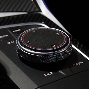 May include: A circular black car infotainment control knob with a textured metallic silver edge. The top surface features four white directional arrows and a thin red and blue circular accent line. Surrounding buttons are labeled HOME, MEDIA, COM, BACK, and OPTION.