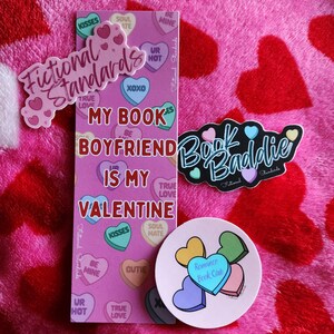 May include: A pink and red heart-patterned blanket with a variety of stickers on it. The stickers are all heart-shaped and feature different phrases like "My Book Boyfriend is My Valentine", "Book Baddie", and "Romance Book Club".