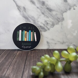 May include: A round sticker with a black background and white text that reads "I'm All Booked". The sticker features a stack of colorful books with green leaves on the spines.