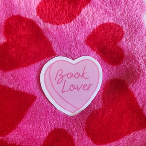 May include: A pink heart-shaped sticker with the text "Book Lover" on a pink and red fuzzy background with hearts.