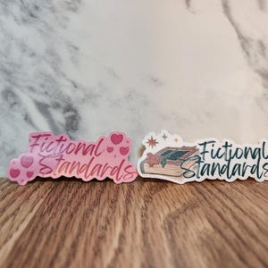 May include: Two stickers with the text "Fictional Standards" in pink and white. The pink sticker has pink hearts and the white sticker has a drawing of a stack of books with a rose.