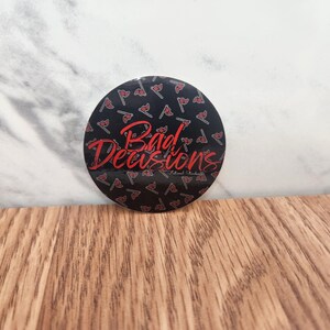May include: A black and red round sticker with a repeating pattern of red axes on a black background. The sticker has the text "Bad Decisions" in red cursive lettering.