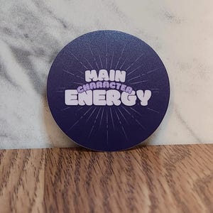 May include: A round sticker with a dark blue background and a white starburst design. The text "MAIN CHARACTER ENERGY" is written in a purple font.