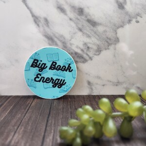 May include: A blue and white round sticker with a book pattern and the text "Big Book Energy" in black.